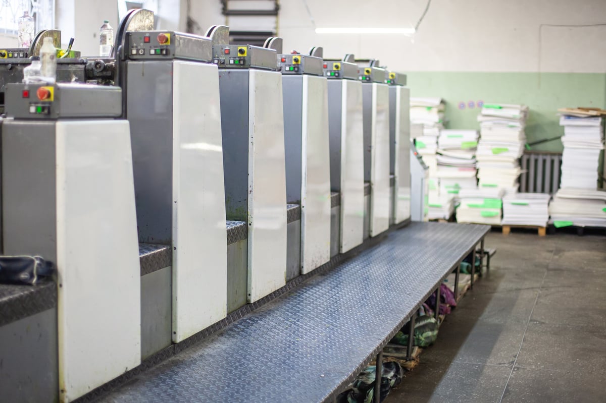 Interior de una imprenta industrial con línea de máquinas de impresión offset y pilas de papel